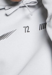 Svetle šedá mikina s jemnou texturou, na přední straně s černými grafikami: číslo "72" a pruhované vzory. Šňůrka u výstřihu.