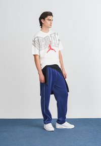 Valkoinen lyhythihainen t-paita, jossa on punainen grafiikka ja mustat yksityiskohdat, yhdistettynä sinisiin verkkareihin. Valkoiset lenkkarit täydentävät asukokonaisuuden.