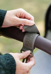 Brun læderhåndtag med et gråt stofbånd sikret med en justerbar løkke. En hånd fastgør båndet til håndtaget.