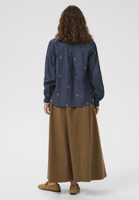 Sötétkék farmering hímzett pillangómintákkal, egy barna, földig érő szoknyával párosítva. Az öltözet hosszú ujjú és laza fazonú.