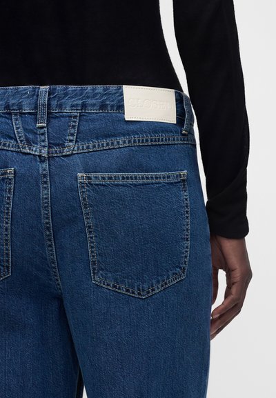 Jean en denim bleu foncé avec poches arrière et un patch de marque en cuir blanc marqué "CLOSED", porté avec un haut noir à manches longues.