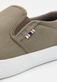 Olivově zelená plátěná slip-on obuv s texturovaným svrškem, hnědým elastickým detailem a malým pruhovaným akcentem v červené, bílé a modré barvě na boku.