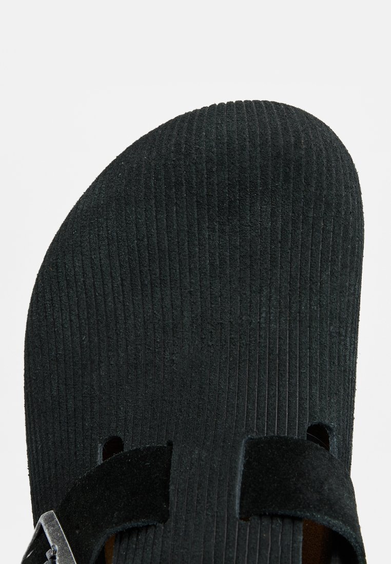 Schwarze Wildledersandale mit gerippter Textur, ausgestattet mit zwei Riemen und einer silbernen Schnalle. Offene Zehenpartie mit ergonomisch geformtem Fußbett für zusätzlichen Komfort.