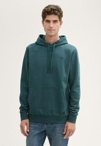 Felpa verde con cappuccio in un tessuto morbido e texture, dotata di tasca frontale e coulisse regolabili, indossata con jeans in denim blu.