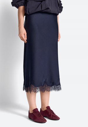 Isik, kes kannab tumesinist keskmise pikkusega seelikut pitsilise äärega ja burgundia värvi tossusid, seisab ühevärvilise heleda tausta ees.