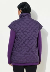 Lila gesteppte Weste mit hohem Kragen und kurzen Ärmeln, die mit strukturiertem Stich verziert ist. Tragen über einem dunkler lilanen Langarmshirt.