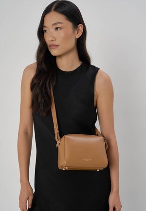 Femme aux longs cheveux bruns portant une robe noire sans manches et un petit sac à bandoulière en cuir beige.