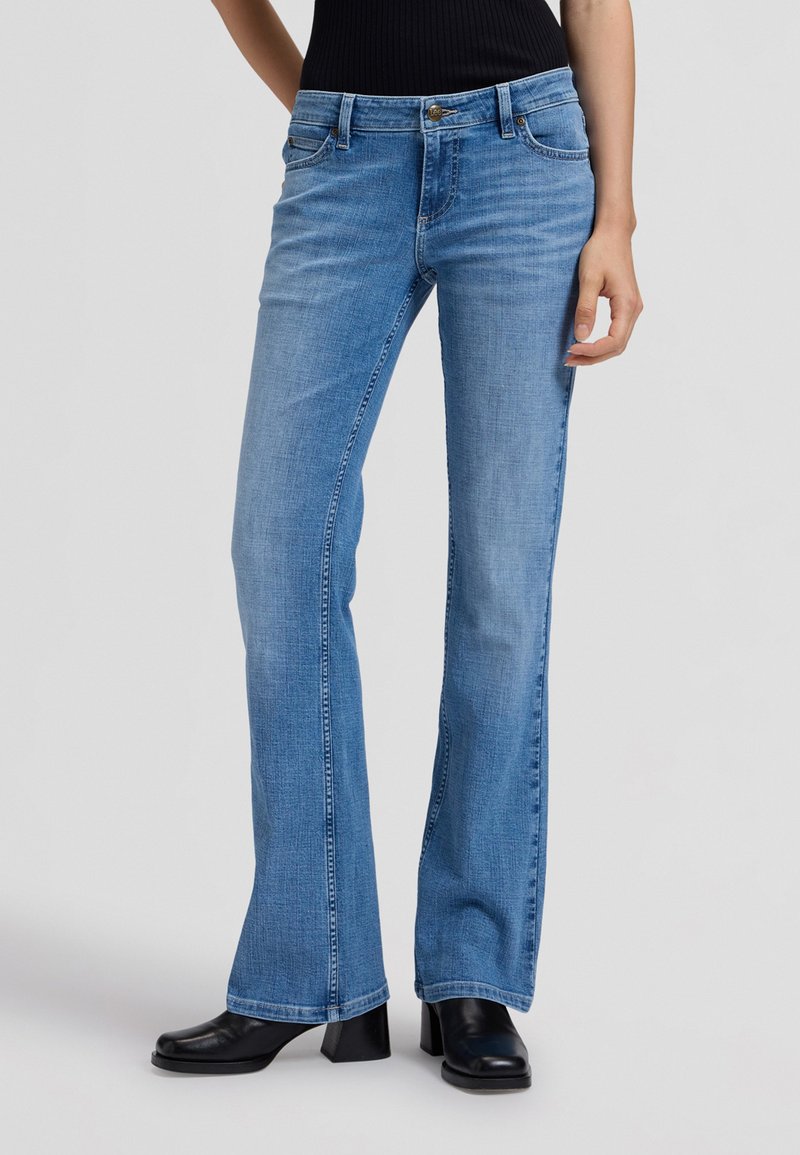 Femme portant un jean bootcut bleu clair et des bottines noires à talons, debout devant un fond gris uni.