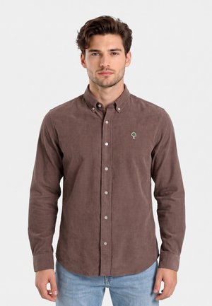 Uomo che indossa una camicia marrone con bottoni e un piccolo logo verde a forma di albero sul petto e jeans azzurri, in piedi davanti a uno sfondo bianco.