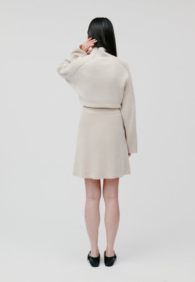 Beige gerippter Pullover mit hohem Kragen, kombiniert mit einem passenden gerippten Rock mit ausgestelltem Saum. Dazu werden schwarze Schuhe getragen.