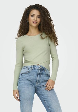Hellgrünes, geripptes Langarmoberteil mit Wickeldesign, kombiniert mit hochgeschnittenen hellblauen Jeans. Glatte Textur, figurbetonte Passform.