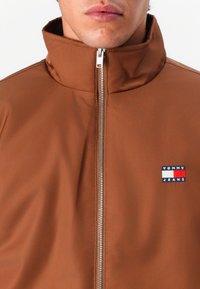 Brun lynlåsjakke med høj krave og et lille Tommy Jeans-logo i blå, rød og hvid på venstre bryst.