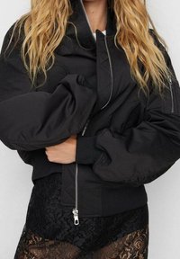 Blouson aviateur noir avec un col montant et des manches surdimensionnées, doté d'une fermeture éclair et de poignets côtelés. Dentelle noire texturée en dessous.