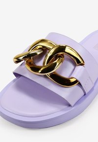Sandalia deslizante de lavanda con una banda ancha que presenta acentos de cadena en tono dorado entrelazados. Textura suave, suela plana y diseño minimalista.