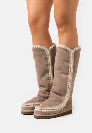 Knee-high brown suede boots with a textured finish, featuring a contrasting cream braided trim and a flat rubber sole.