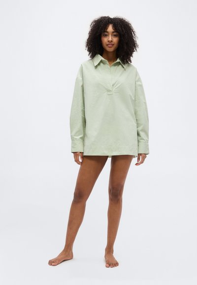 Γυναίκα με σγουρά μαλλιά που στέκεται ξυπόλητη, φορώντας ένα φαρδύ ανοιχτοπράσινο πουκάμισο με μακριά μανίκια, μπροστά σε μονόχρωμο φόντο.