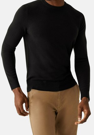 Homme portant un pull noir ajusté à manches longues et un pantalon sable, debout avec les bras détendus le long du corps sur un fond uni.