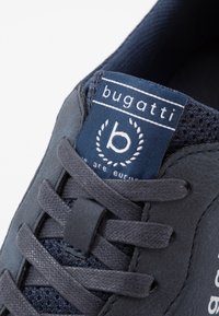 Navy sneaker with suede and mesh materials, featuring flat laces, a padded collar, and a branded tongue label with a logo.