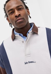 Πολυκαναλισμένο πόλο με μπεζ σώμα, ναυτική μπλε μανίκια και καφέ γιακά. Διαθέτει κεντημένο κείμενο "iets francs..." στο στήθος.