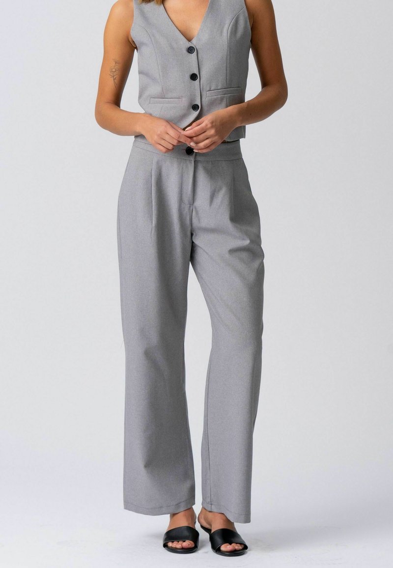 Femme portant un gilet gris clair boutonné et un pantalon large assorti, debout les mains jointes, chaussée de sandales à glissière noires à bout ouvert.