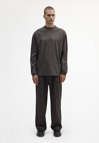 Langärmliges, dunkelgrünes Baumwoll-T-Shirt mit Rundhalsausschnitt, kombiniert mit lockeren, dunkelbraunen Hosen und schwarzen Schuhen, vor einem schlichten weißen Hintergrund.