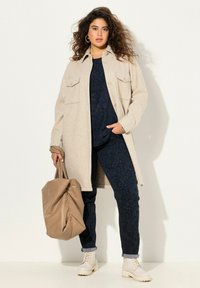 Béžový oversized kabát s předními kapsami, přes námořnický vzorovaný top, námořnické vzorované kalhoty a krémové combat boty, držící béžovou tašku.