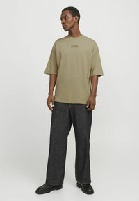 Camiseta oversize de color verde oliva con mangas cortas, que presenta una impresión de texto negro en el pecho. Combinada con unos vaqueros de pierna ancha de color azul oscuro y zapatos negros.