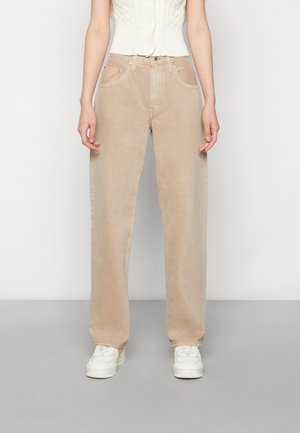 Person, die hoch taillierte beige Jeans mit weitem Bein, weißes ärmelloses Stricktop und weiße Turnschuhe trägt, steht vor einem einfarbigen Hintergrund.