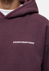 Bordowa bluza z kapturem wykonana z miękkiego materiału, z białym haftowanym logo "GOODFORNOTHING" po lewej stronie klatki piersiowej. Regulowany kaptur.