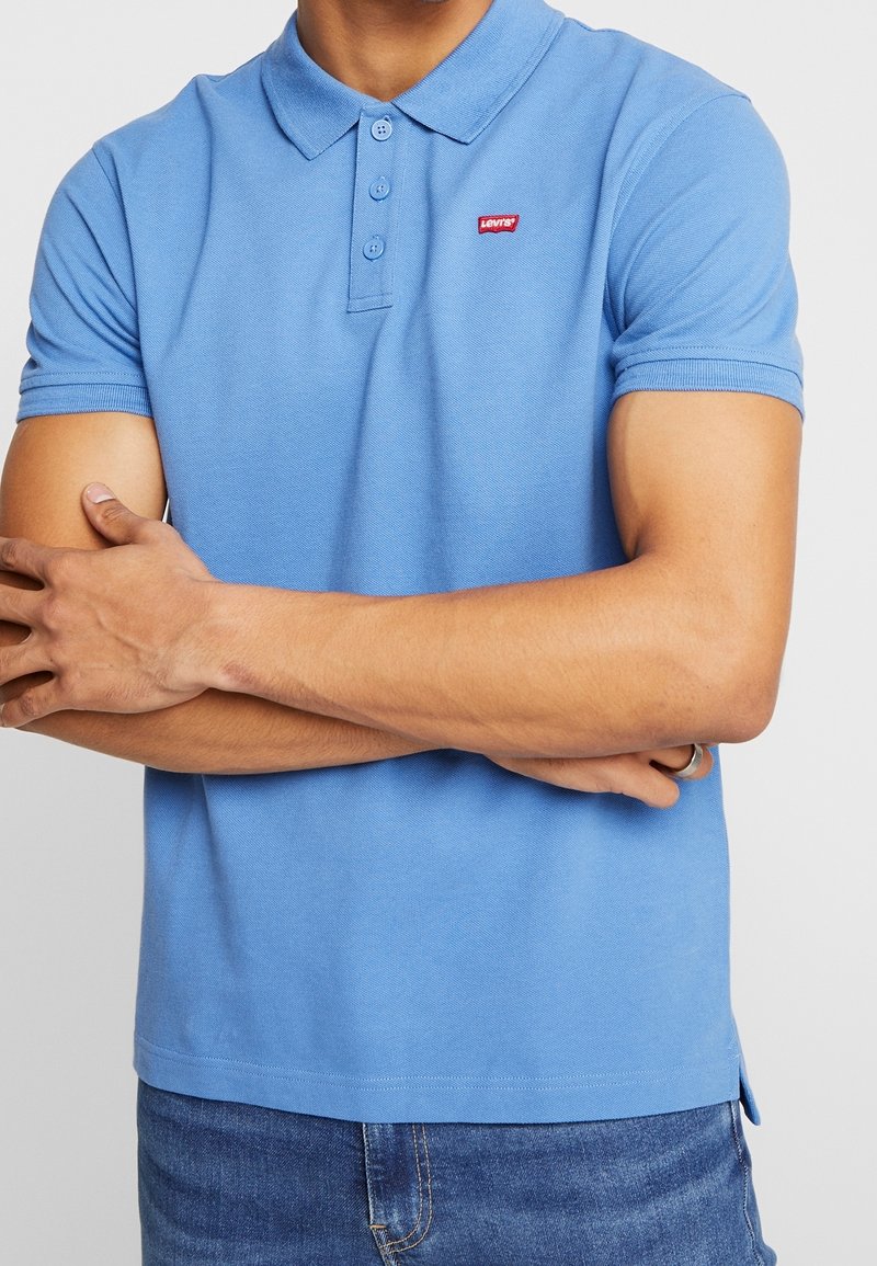 Polo bleu clair en tissu texturé, doté d'un col et d'une patte de boutonnage à trois boutons, avec un petit logo rouge sur le côté gauche de la poitrine.