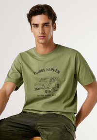 Camiseta de algodón verde oliva con un estampado gráfico negro de una furgoneta y texto que dice "LAS COSAS SUCEDEN, VA A LA DISTANCIA EXTRA" en el frente.