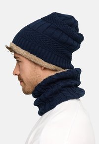 Navy knitted beanie with tan faux fur trim and matching neck gaiter, featuring ribbed and cable patterns for added texture and warmth.