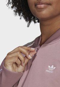 Chaqueta deportiva morada con cremallera y puños acanalados, con detalles de tres rayas blancas y un logo bordado en el pecho izquierdo.