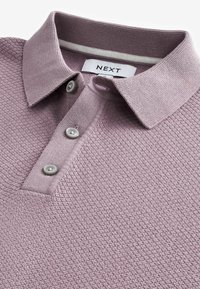 Vaalean lilac polo-paita, jossa on teksturoitu kangas, kaksi nappia ja ribattu kaulus, jossa on valkoinen raita. Tunnuksessa lukee "NEXT."
