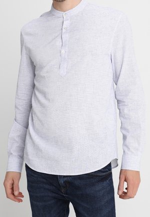 Homme portant un shirt henley à manches longues gris clair avec trois boutons et un jean bleu foncé, debout devant un fond uni.