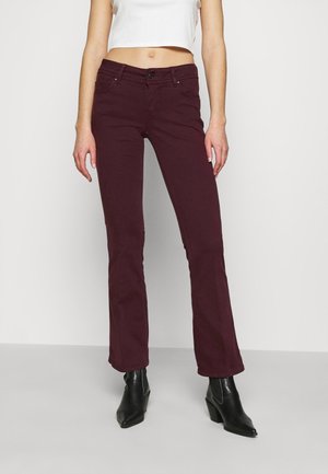 Donna che indossa jeans color borgogna scuro a gamba dritta, top bianco corto e stivaletti neri con punta a punta, in piedi contro uno sfondo semplice.