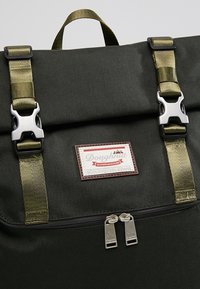 Ciemnozielony plecak wykonany z płótna, z oliwkowymi paskami, srebrnymi elementami, z zamkiem błyskawicznym oraz skórzaną łatą z logo.