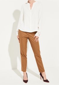 Weiße Langarmbluse mit V-Ausschnitt und Spitze an den Manschetten, kombiniert mit braunen taillierten Hosen und dunkelroten High Heels.