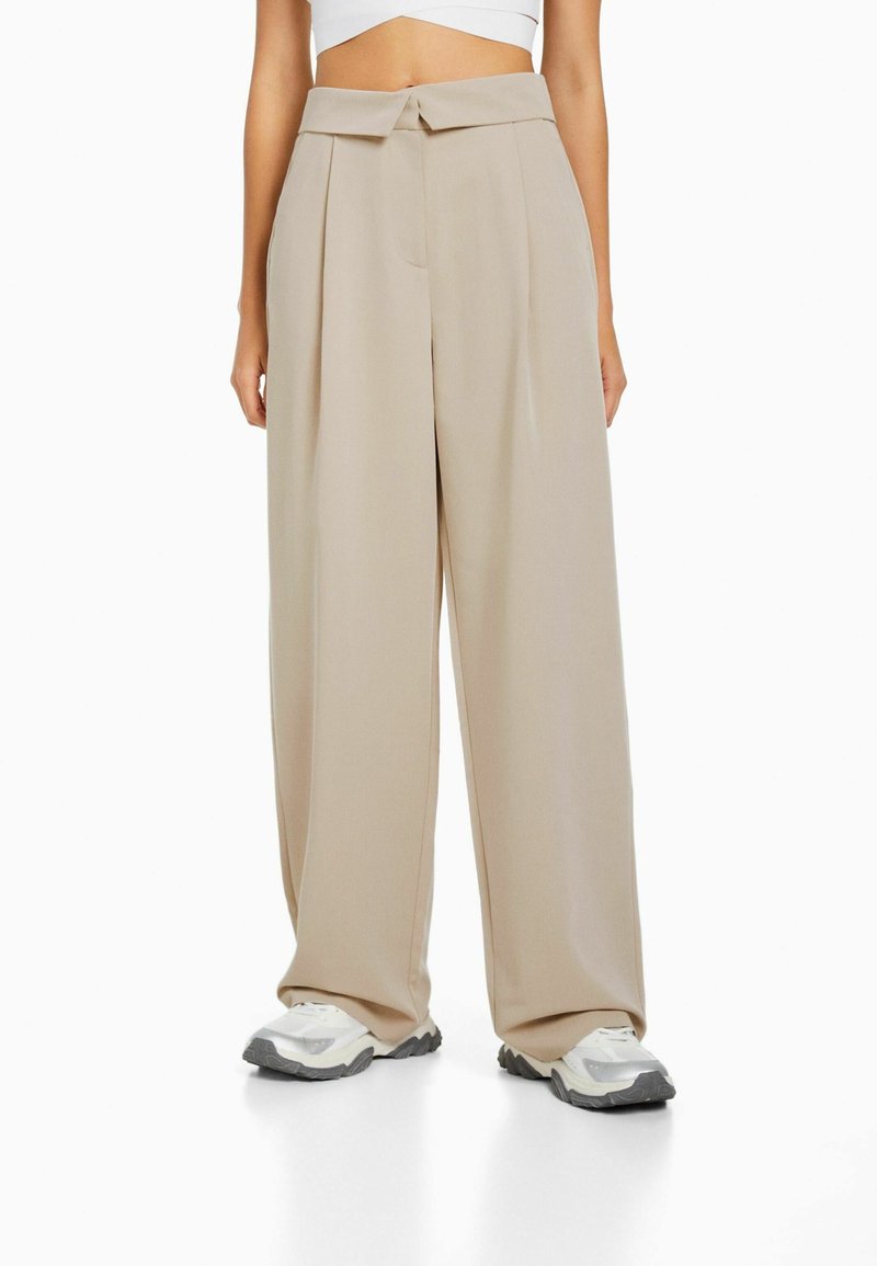 Pantalons larges beiges avec une texture lisse, dotés d'un devant plissé et d'un détail nœud papillon à la taille. Associés à des baskets.