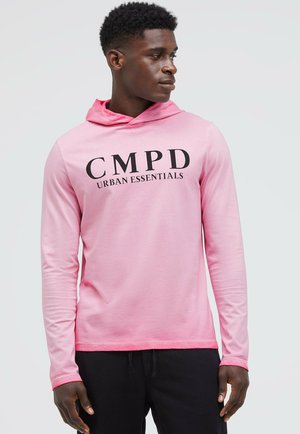 Junger Mann trägt einen langärmligen rosa Hoodie mit dem Text "CMPD Urban Essentials", schaut seitlich vor einem einfachen hellen Hintergrund.
