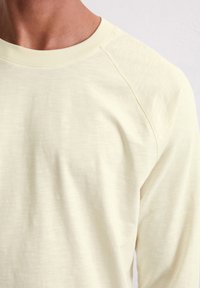 Hellgelbes Langarmshirt mit glatter Struktur und Raglanärmeln. Keine auffälligen Muster oder Hardware sichtbar.