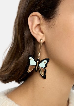Zbliżenie na ucho kobiety noszącej złoto-czarną, motylowatą kolczyk wiszący z jasnoniebieskimi i metalicznie złotymi detalami.