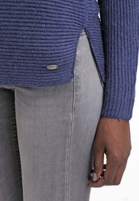 Pull en maille côtelée Khujo bleu foncé, avec un ourlet arrondi et des fentes latérales. Associé à un jean ajusté gris clair et un détail logo argenté.