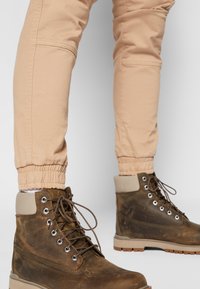 Bottes en cuir marron à la cheville avec une finition mate, œillets en métal et lacets marron. Associées à un pantalon beige fuselé avec des bas élastiques.