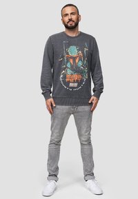 Sudadera gris oscuro con un gráfico de Boba Fett, que presenta un cuello redondo y un dobladillo acanalado. Combinada con jeans gris claro y zapatillas blancas.