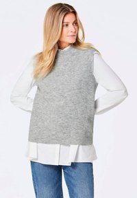 Grauer gestrickter Pullunder mit hohem Kragen, der über einem weißen Hemd mit Kragen getragen wird. Jeans sind darunter sichtbar. Glatte Textur mit dezentem Muster.
