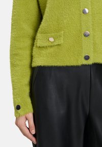 Gros plan sur une personne portant un cardigan moelleux vert lime avec des boutons en argent et un pantalon en cuir noir, la main relâchée sur le côté.