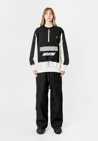 Chaqueta con cremallera negra y crema con un bolsillo en la parte frontal, con acentos grises y el logotipo "MWM". Combinada con pantalones sueltos negros de textura.