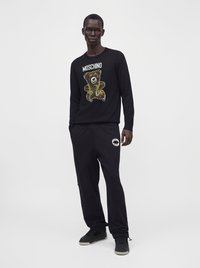 Homme portant un sweat-shirt noir Moschino avec un graphique d'ours en peluche et un pantalon de survêtement noir assorti avec un logo blanc Moschino sur la cuisse gauche.