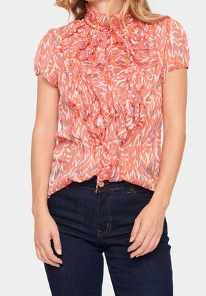 Bluzka w kolorze koralowym z krótkim rękawem i falbaniastym przodem; ma wysoki kołnierz, wzorzystą teksturę i subtelne niebieskie akcenty. Połączona z ciemnymi dżinsami.